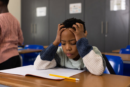 Child and youth classmates sitting at classroom desk, holding head over notebook with yellow pencil