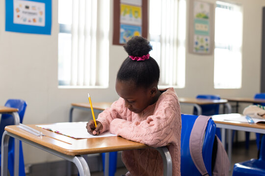 African American child girl writing with yellow pencil on paper at school desk wearing pink sweater