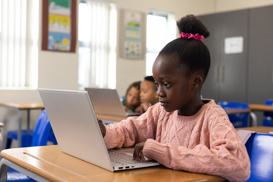 African schoolgirl in pink sweater sitting at wooden desk using silver laptop at school with blinds