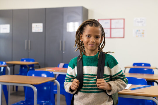 African teen male pupil standing holding backpack straps in classroom wearing green striped sweater