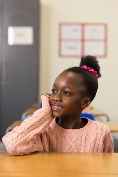 School-age African girl sitting at wooden desk in classroom, wearing pink sweater and scrunchie