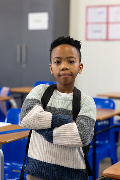 African American boy standing with arms crossed wearing striped sweater and backpack in classroom