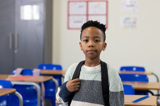 African American child standing holding backpack facing lens in class with desks blue chairs folder