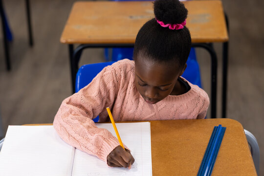 Child writing in lined notebook at classroom desk in pink sweater using yellow pencil