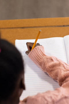 African female child writing in ruled notebook at school desk, pink sleeve holding yellow pencil