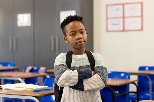 African American boy standing with arms crossed in classroom wearing striped sweater and backpack