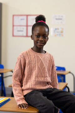 African girl child sitting on wooden desk at school wearing pink sweater, holding colorful folder