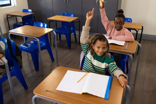 African American child classmates raising hands sitting in classroom at wooden desks with notebooks