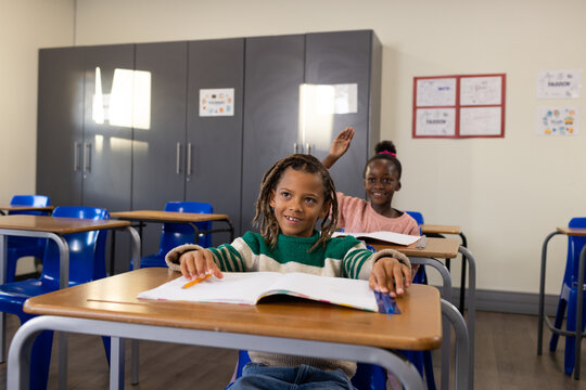Children sitting in classroom at wood desks, blue chairs, front boy writing with pencil in workbook