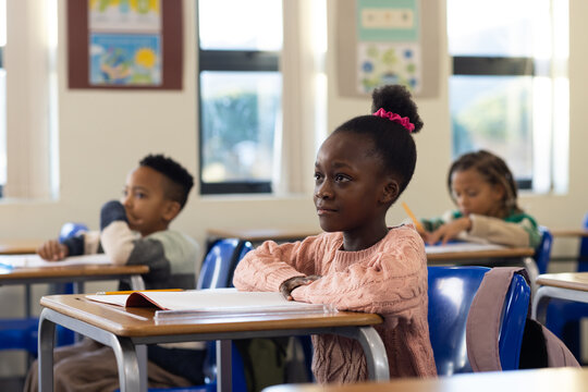 Classmates sitting at desks in classroom, African American girl in pink sweater writing notebook