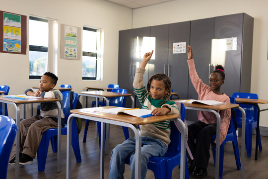 African American students sitting at desks in primary school classroom raising hands with notebooks