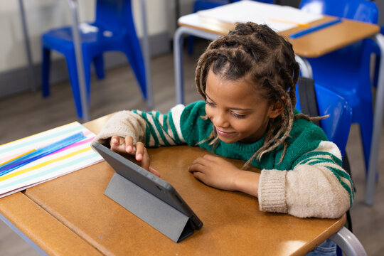 African American male child sitting at school desk wearing green sweater, interacting with tablet