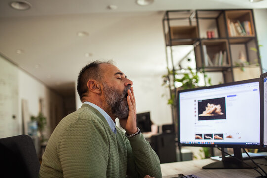 Tired bearded man yawning at computer in modern office workspace
