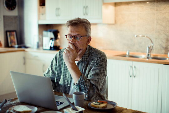 Senior man thinking while working on laptop in home kitchen