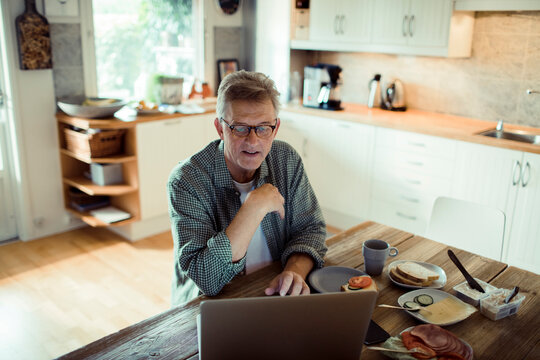 Older man using laptop while having breakfast in home kitchen