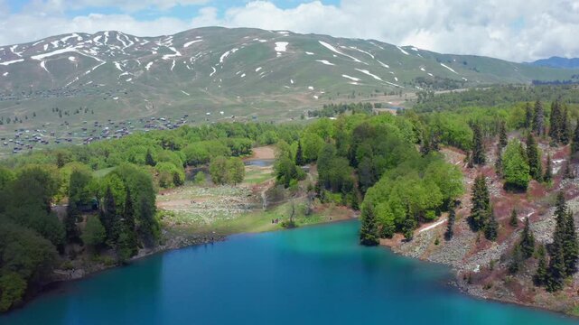 Aerial drone flies forward above Green Lake near Goderdzi in Georgia, framing turquoise water, wooded slopes and lingering snow for nature tourism, hiking media and regional travel campaigns.