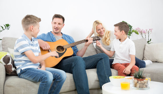 A heartwarming lifestyle shot of a happy family sitting together on a modern sofa in their living room. The father plays an acoustic guitar while his wife and two young sons smile and listen