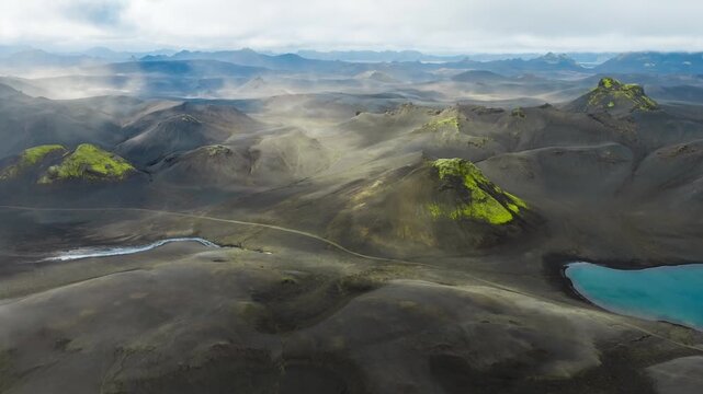 Aerial timelapse over central Iceland highlands shows ash hills, moss craters, turquoise lake, sinuous stream, and a faint track as mist and clouds drift while camera glides forward.