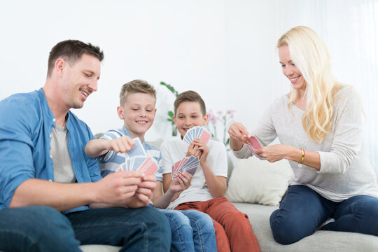 A cheerful lifestyle photo of a family of four sitting on a couch in their living room, playing a card game together. The mother, father, and two young sons are holding playing cards