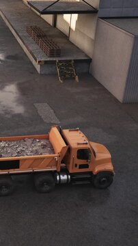 A truck is parked at a construction site. It carries building materials in its bed. Workers move bricks on the ground. The sun is shining and the area is busy with activity.