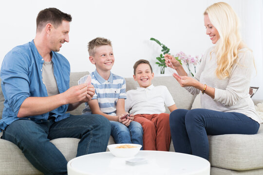A cheerful lifestyle photo of a family of four sitting on a couch in their living room, playing a card game together. The mother, father, and two young sons are holding playing cards