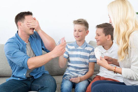 A cheerful lifestyle photo of a family of four sitting on a couch in their living room, playing a card game together. The mother, father, and two young sons are holding playing cards