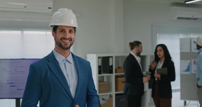 Portrait of Latin American man building company CEO smiles in conference hall. Bearded Hispanic engineer in helmet stands against colleagues in corporate office