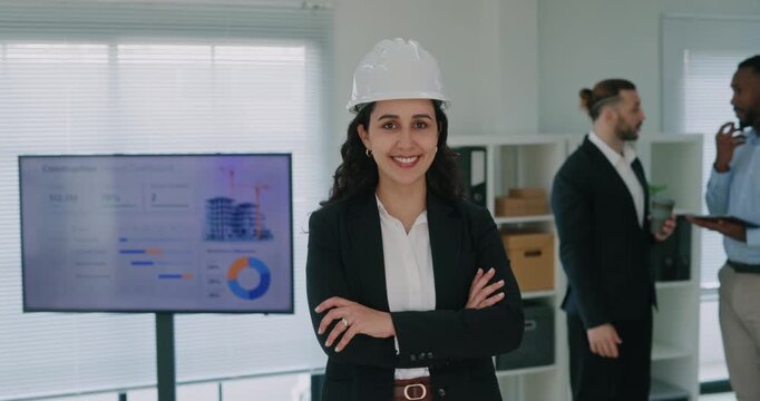 Portrait of Latin American woman construction site manager smiles in conference hall. Hispanic lady architect designer in helmet stands by colleagues in office