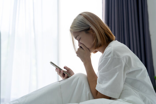Asian woman feels sad looking at her smartphone, feeling depressed after receiving a message, sitting on her bed in her bedroom at home.