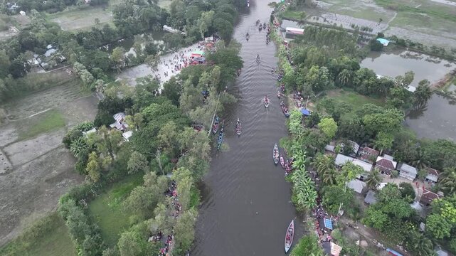 Gopalganj, Bangladesh - 30 October 2023: Aerial view of a traditional boat race on a river, with narrow wooden boats navigating the waterway surrounded by lush greenery and rural homes.