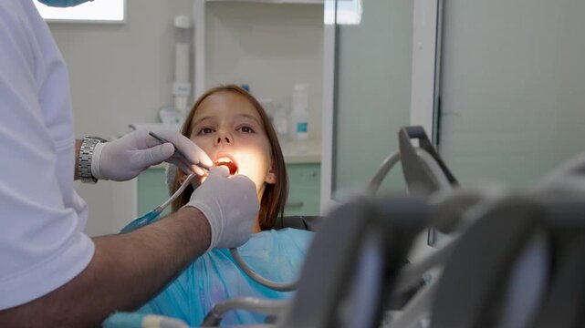 Pediatric dentist examining teeth of young girl sitting in dental chair.