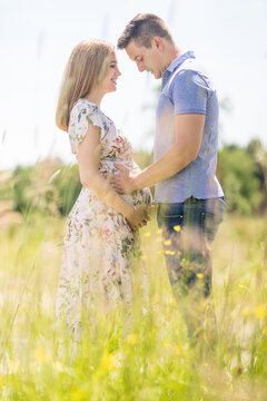 Side view of a happy pregnant couple embracing gently and holding the belly in a sunlit grassy field during summer. Romantic maternity concept of love, family preparation, and new life.