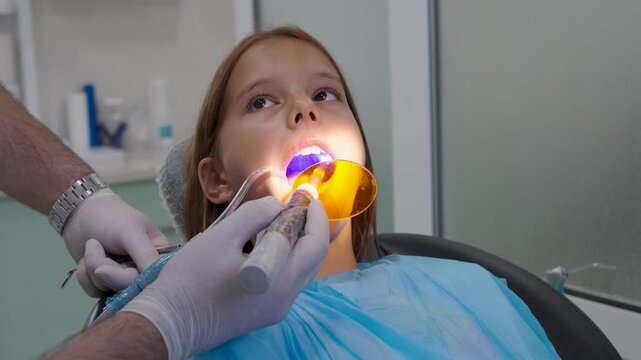 Pediatric dentist using UV curing light lamp on tooth filling for young girl.