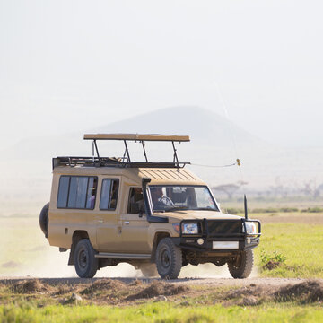 A beige 4x4 safari truck with a raised pop-up roof driving along a dirt road through the open grassy savanna plains of an African national park during a game drive excursion.