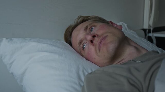 Young adult Caucasian man lying in bed awake, staring at ceiling with worried expression, suffering from insomnia. Video for mental health advocacy or sleep disorder treatment promotions
