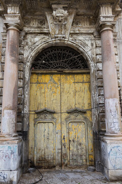 An ornate vintage yellow wooden entrance door framed by stone columns and an arched doorway featuring a carved grotesque mask keystone and intricate iron fanlight transom.