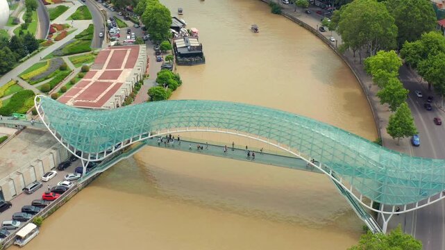 Aerial drone descends toward the Bridge of Peace in Tbilisi, Georgia, revealing its glass canopy, Kura River and Rike Park for architecture tourism, city travel and landmark promotion campaigns.