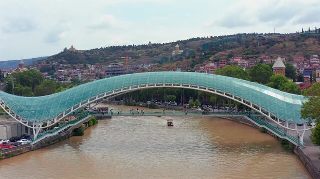 Aerial drone retreats from the Bridge of Peace in Tbilisi, Georgia, widening across the glass structure, Kura River and hillside cityscape for architecture, tourism and destination advertising.