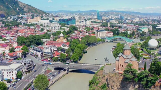 Metekhi Church anchors circling aerial view above the river junction, historic rooftops and central Tbilisi streets, supporting cultural tourism, city mapping and Georgian heritage destination media.