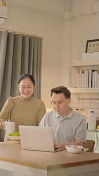 Senior couple enjoying peaceful morning together at home. Elderly Asian man and woman sit together smiling and talking with laptop and breakfast on the table. healthy aging, retired lifestyle.