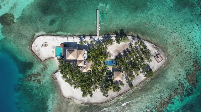 Utila, Honduras - 02 April 2023: Aerial view of a private island resort featuring palm trees, a swimming pool, a tennis court, and a wooden pier surrounded by clear turquoise Caribbean waters.