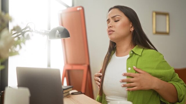 Woman at laptop clutching neck with hand while stretching opposite arm beside desk lamp and mug in building with books and window visible; neck pain.