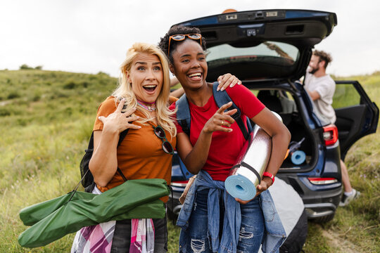 Two cheerful women stand by their car, excited for a camping trip. They wear casual outfits and hold camping gear, surrounded by grassy hills under bright sunlight.