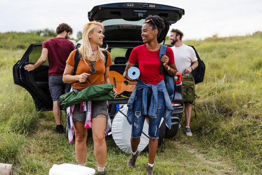 Two friends carry camping supplies from their vehicle, smiling and enjoying the moment amidst a lush green landscape. A cheerful atmosphere surrounds them.