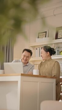 Senior couple enjoying peaceful morning together at home. Elderly Asian man and woman sit together smiling and talking with laptop and breakfast on the table. healthy aging, retired lifestyle.