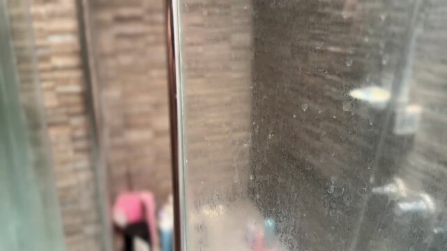Closeup of a dirty shower glass door with white mineral residue from hard water and soap scum. Blurred shower cabin in the background. Common bathroom cleaning issue.