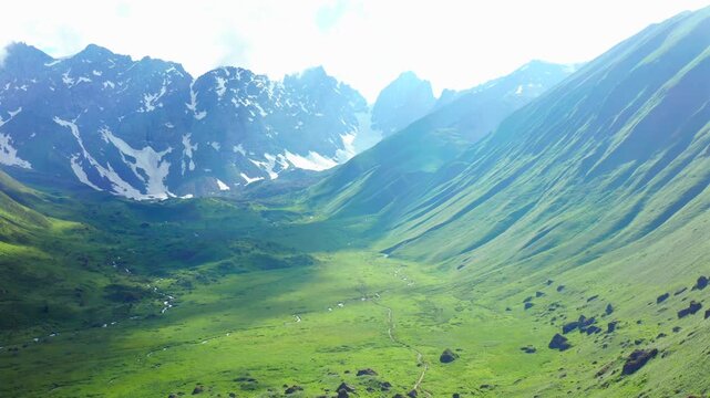 Drone descends above an alpine valley near Roshka, Georgia, revealing Chaukhi peaks, green meadows and snowmelt streams for trekking tourism, hiking promotion and mountain destination media.