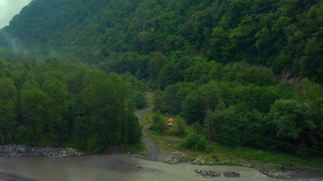 Drone retreats above an Aragvi riverside clearing in Georgia, revealing a small orange tent, forest edge and mountain river for camping promotion, wilderness travel and outdoor recreation media.