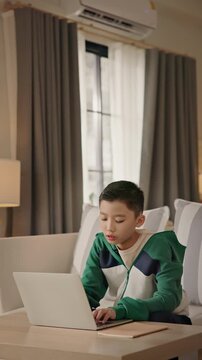 A young Asian boy sits comfortably at home using a laptop for gaming, studying, or browsing online. Modern childhood, digital education, balance of learning and leisure through technology. Gen Alpha.