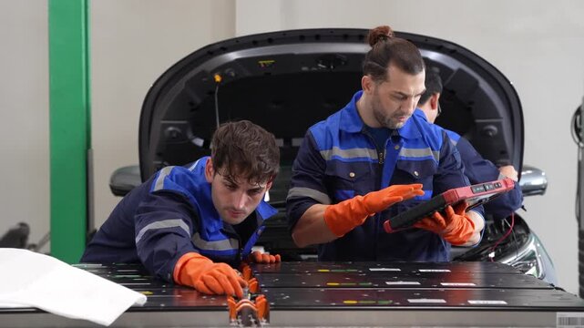Mechanic technician electric vehicle battery diagnostic workshop maintenance safety, team in uniform inspecting high voltage battery pack under open hood, using tablet scan tool, careful repair